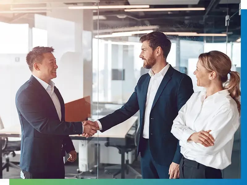 Asiatisch und europäisch gelesene Personen geben sich in einem hellen Büro die Hand als Symbol als globale Wirtschaftsbeziehungen durch SIBA GLOBAL